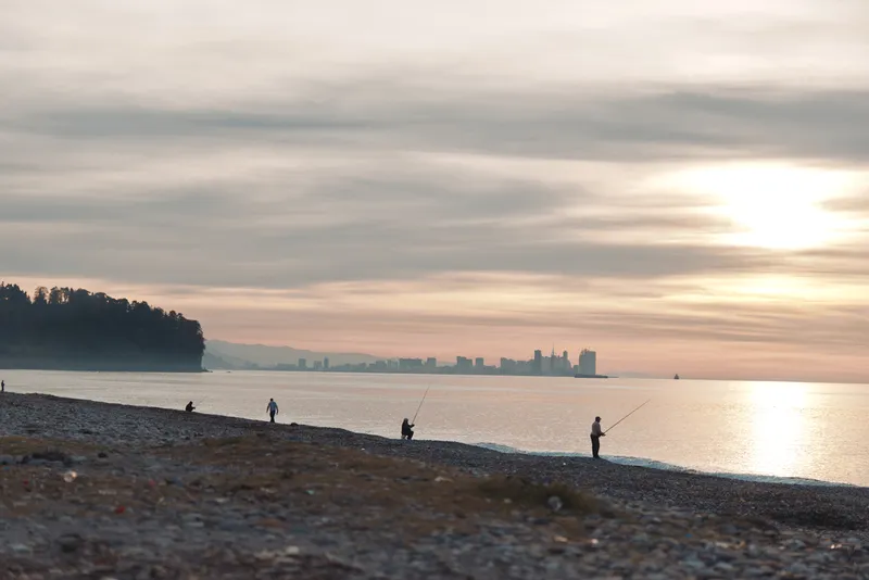 Beach at sunset with silhouettes of fishermen along the calm shore, water reflecting warm light, pebbles scattered on sand, and a tranquil horizon over a quiet coastal scene