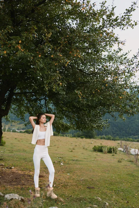 Young woman in white outfit stands relaxed under a large tree in a green meadow enjoying a peaceful moment outdoors.