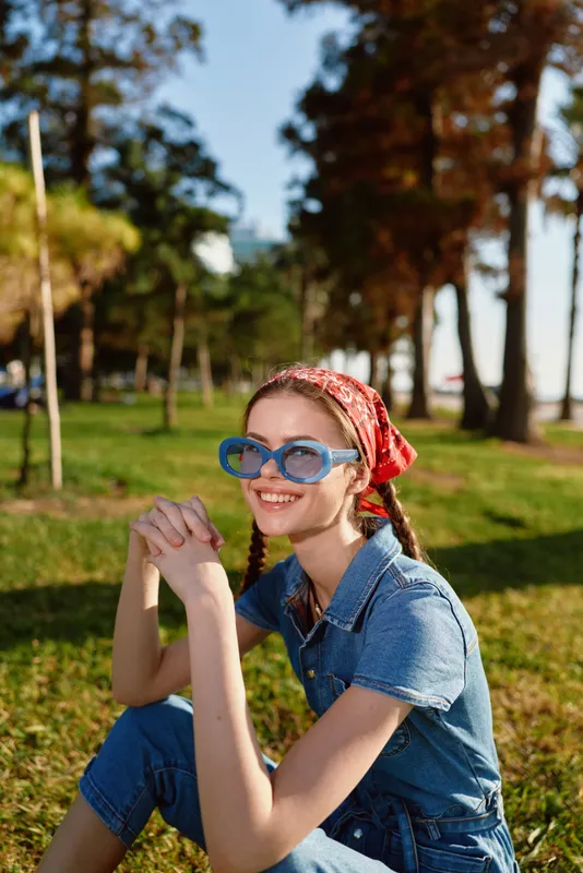 Smiling woman wearing blue sunglasses and red headscarf sits on green grass outdoors in sunny park with trees and warm natural light