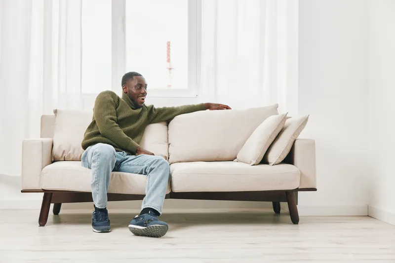 Comfortable young Black man relaxing on a beige sofa in a bright and modern living room, wearing casual clothing, with a calm and satisfied expression