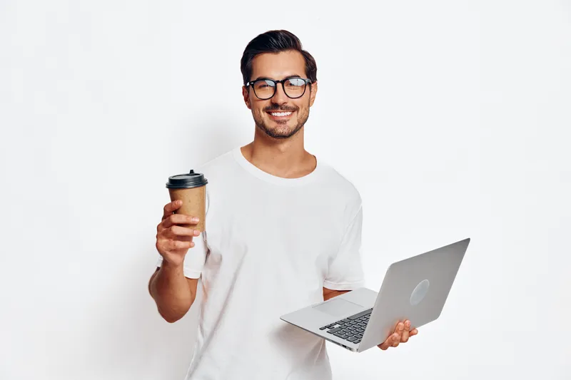 Smiling young man wearing glasses holds a laptop in one hand and a coffee cup in the other, exuding a sense of productivity and happiness against a white background.