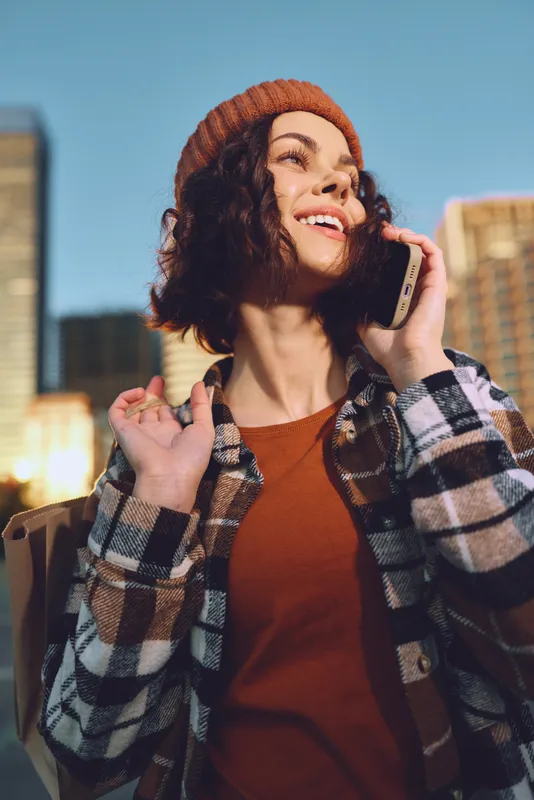 woman phone smile city candid lifestyle golden beanie authenticity glow emotional storytelling mindful living: Young woman in beanie smiles on phone in city at golden hour, authentic urban moment.