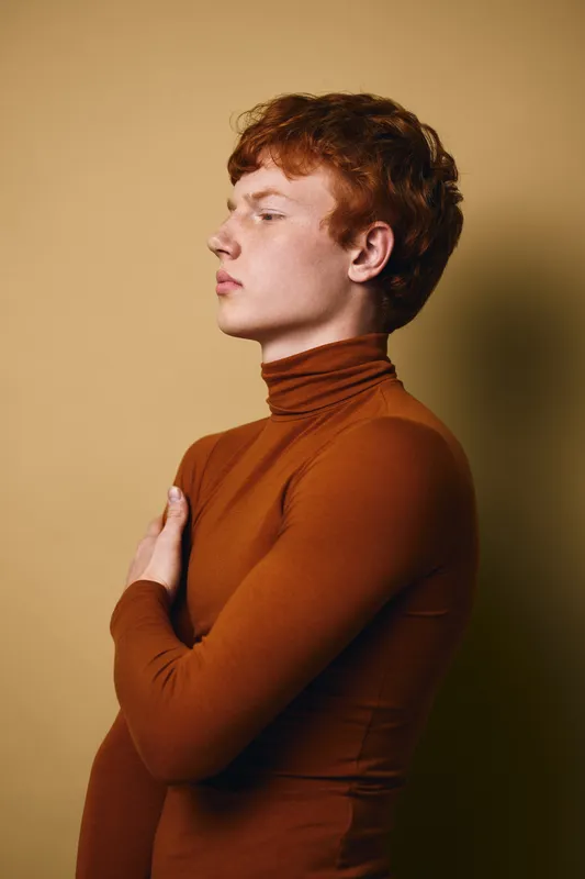 Young man with red hair and freckles in brown turtleneck poses with arm across chest against beige background in profile studio portrait with serious expression.