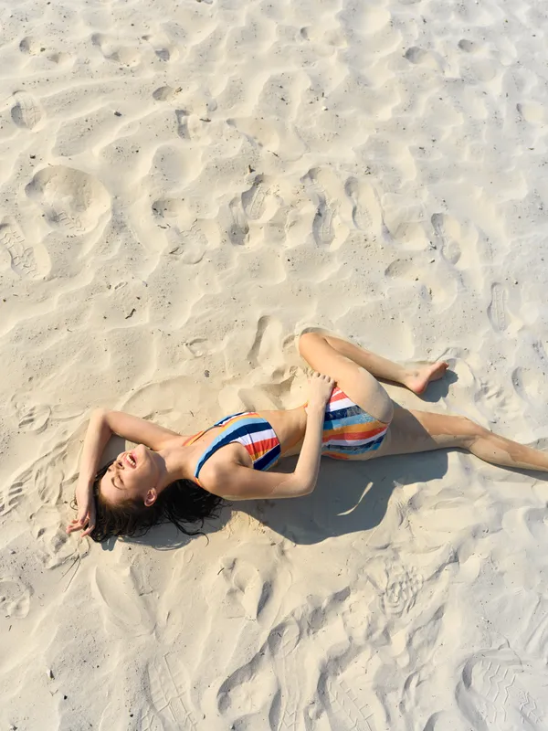 A relaxed woman lies on sunlit sand wearing a striped bikini, basking in warm light and gentle breeze for a carefree seaside moment.