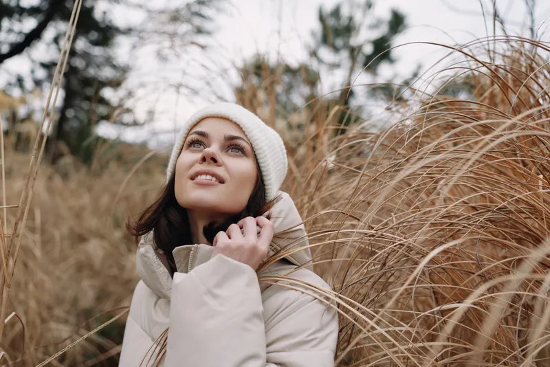 Woman in a light coat and knit beanie stands among tall autumn grass, looking up with a gentle smile. Soft natural light highlights texture and the cozy outdoor mood with calm atmosphere