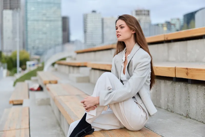 Business woman sitting thoughtfully on urban wooden bench dressed in beige suit with modern city skyline background in natural light KEYWORDS: business woman, urban, city, suit, thoughtful, modern, ou