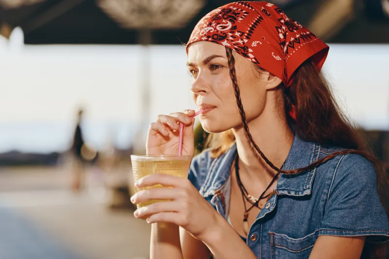 Lifestyle woman in modern boho-western denim street style drinking a cold beverage outdoors with a red bandana, natural light, and warm film color tones.