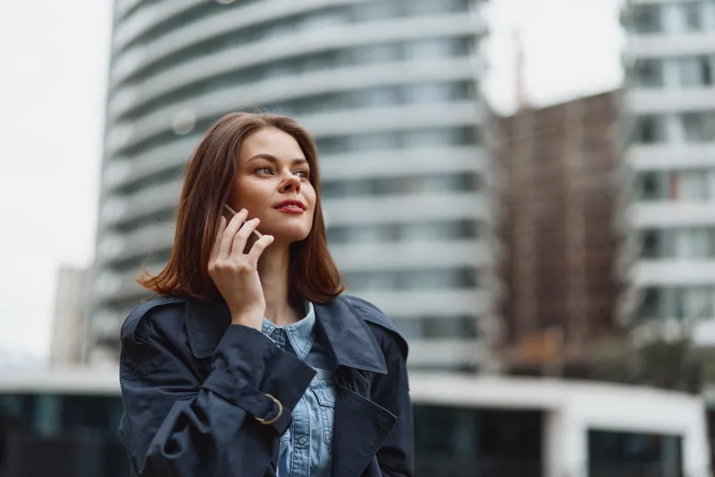 Young woman with brown hair wearing a denim shirt and dark jacket talks on the phone while standing outdoors in an urban setting with modern buildings behind her.