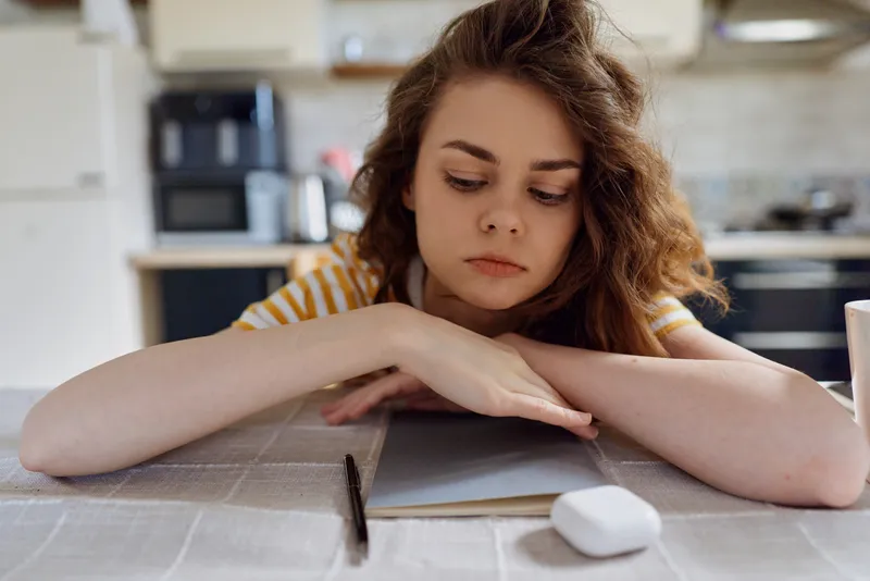 A young woman rests her head on a kitchen table beside a notebook and pencil, conveying calm solitary mood and thoughtful home life in a quiet domestic setting.