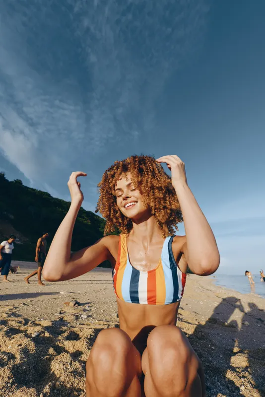 Smiling woman with curly hair wearing a striped swimsuit, relaxing on beach sand, sunny summer day, blue sky background, outdoor vacation feeling, natural light, carefree pose, relaxed
