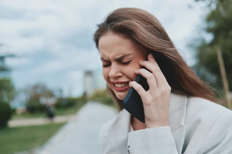 Stressed young woman in light blazer experiencing financial crisis talking on smartphone outdoors with cloudy sky background expressing anxiety and frustration