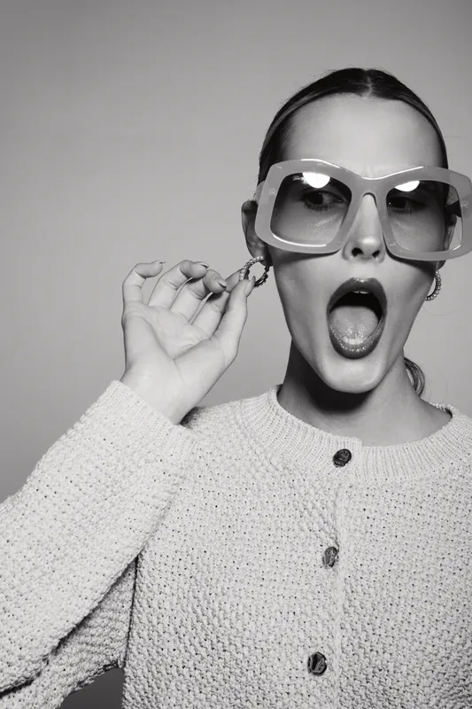 Stylish Caucasian woman with light skin wearing oversized sunglasses and knitted sweater shows surprised expression.