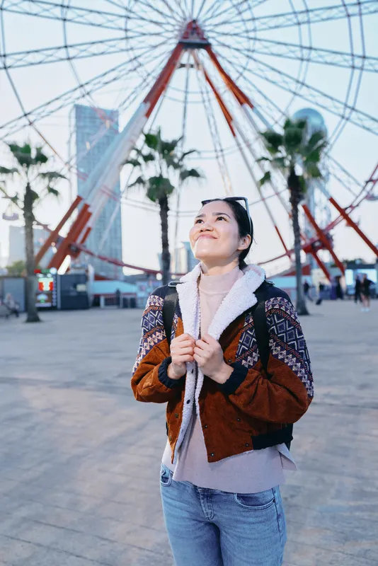 Ferris wheel in the background as a woman stands outdoors wearing a cozy jacket and sweater, looking up with a smile, palm trees and city skyline around, playful adventure
