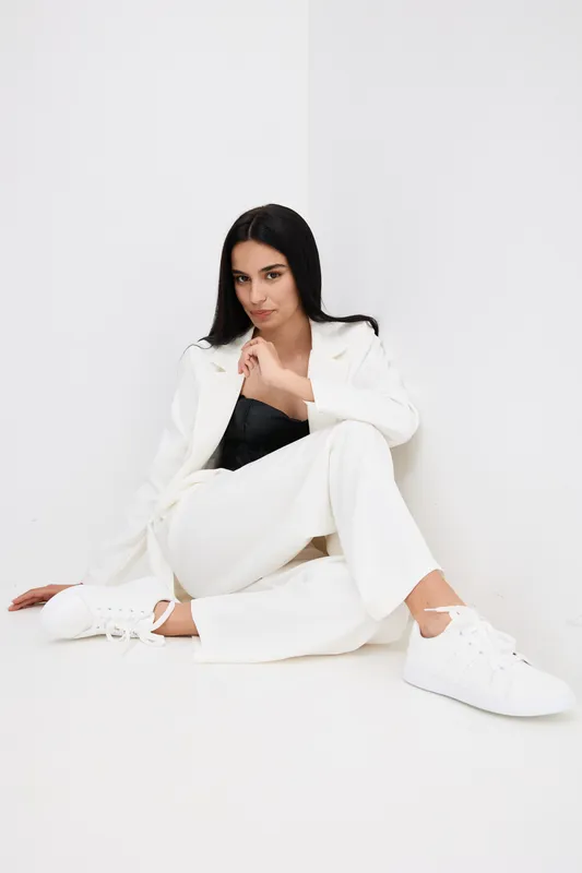 stylish woman in white pantsuit and sneakers sits casually with confident pose against a plain white background, showcasing modern fashion and minimalistic style
