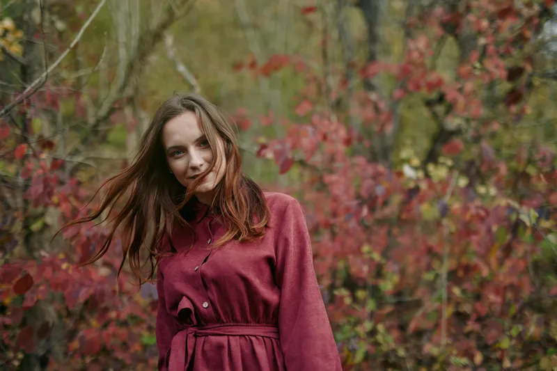 Authentic portrait of a woman in a burgundy dress in a natural autumn landscape, soft light, candid pose, honest mood, subtle details, and a credible, timeless feel.