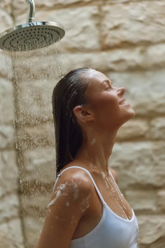 smiling woman enjoying outdoor shower with water droplets and wet hair, wearing white tank top against rustic stone wall background in warm natural light