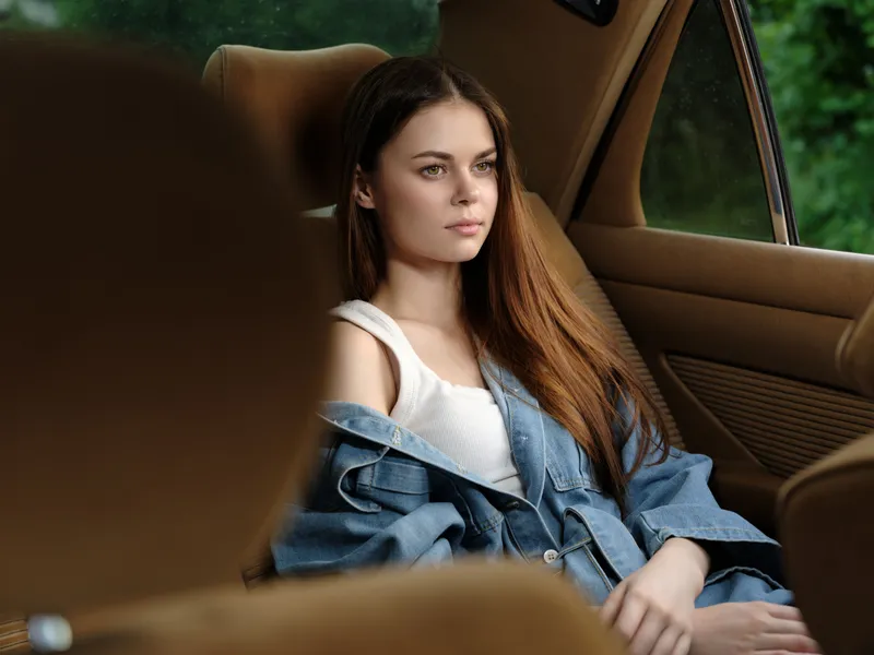 Young woman with long hair sitting in a car, wearing casual denim clothing, gazing thoughtfully out the window, in a serene green setting.