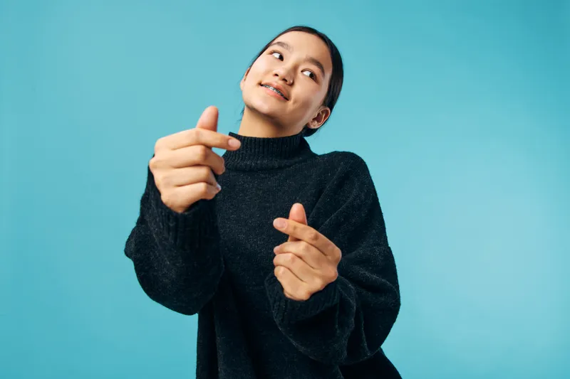 Young woman smiling and making heart gestures with fingers against blue background. Casual clothing, black sweater, emotional expression with positive happy mood in studio.