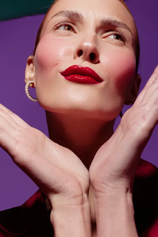 Close-up of a woman with bold red lipstick and pearl earrings posing with hands near face against purple background. Concept of beauty, makeup, fashion, and confident expression.