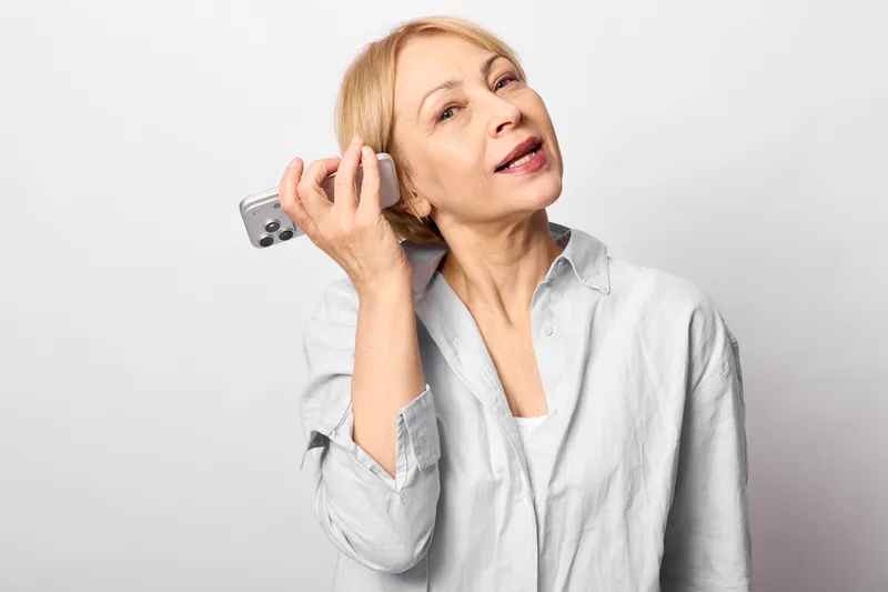 Woman elderly phone conversation stress headache worried indoors white background, she holds a mobile to her ear, tense facial expression shows anxiety, burnout concept