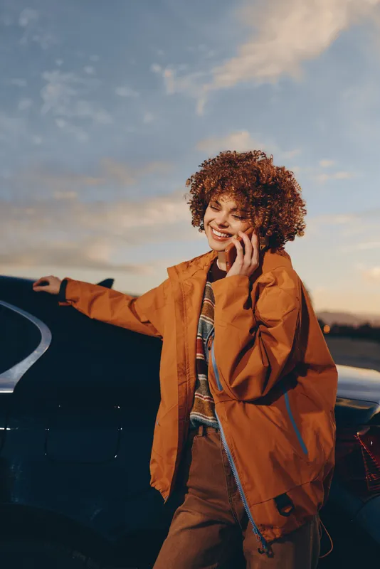 Woman lifestyle, car at sunset, road trip, orange jacket, curly hair, wide smile, outdoor portrait by parked vehicle on highway at golden evening, relaxed fashion mood today
