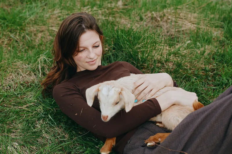 Relaxed woman lying on green grass, cradling a playful goat kid in her arms, outdoor calm and warmth, natural light, gentle lifestyle moment of connection with animals.