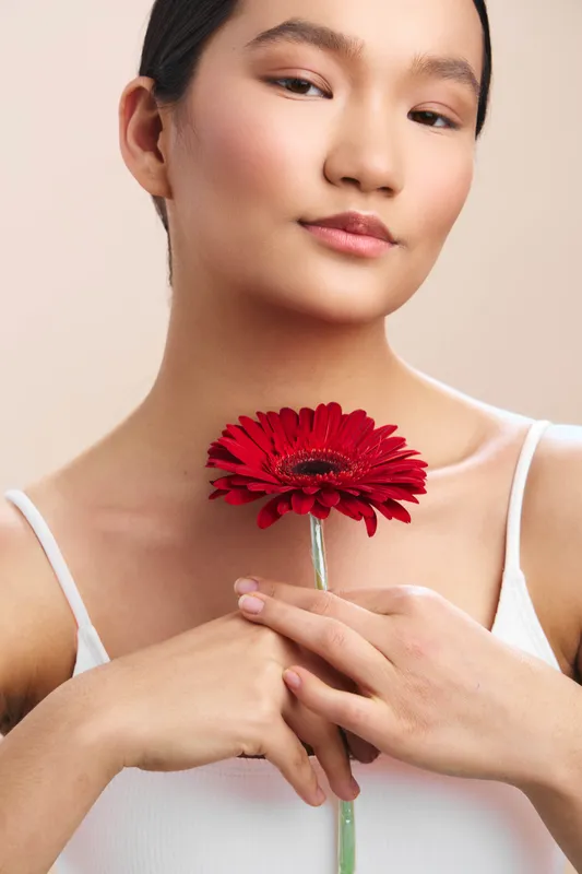 asian woman holds a red gerbera flower close to her chest in a simple studio setting, expressing calm grace and serene beauty that creates a focused, elegant mood.