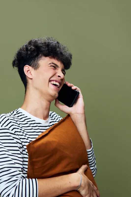 Young man laughing while talking on smartphone, holding a pillow against olive green background, casual striped shirt, joyful and relaxed mood.