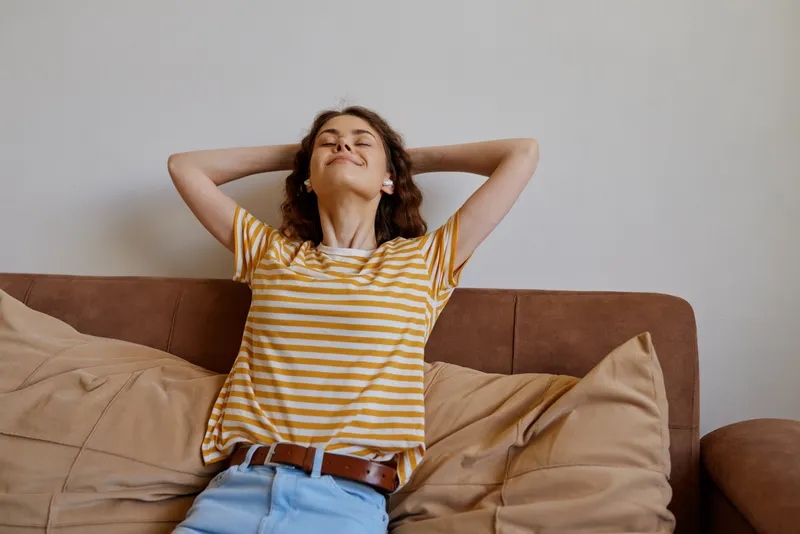 Relaxed woman sits on a couch with arms behind head wearing a striped shirt and jeans, smiling softly. Bright indoor setting conveys casual home lifestyle, comfort, and serene wellbeing. Natural