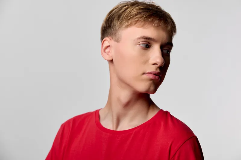 Young man in a red shirt stands against a clean studio backdrop, looking sideways with a calm expression, modern casual fashion vibe, soft lighting, minimal composition for stock use