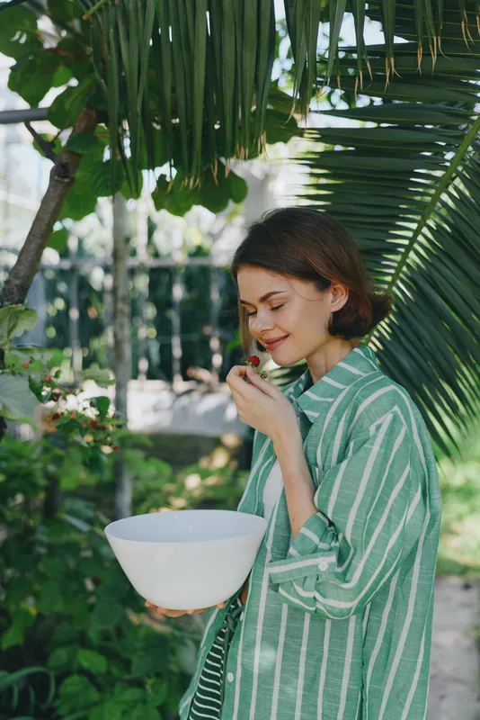 A woman stands in a lush garden wearing a green striped robe, smiling softly while holding a white bowl.