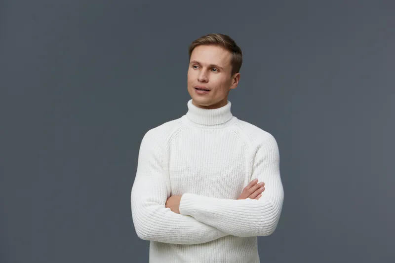 Handsome young man wearing white knitted turtleneck sweater looking thoughtfully to the side isolated on plain grey background studio portrait. People lifestyle concept