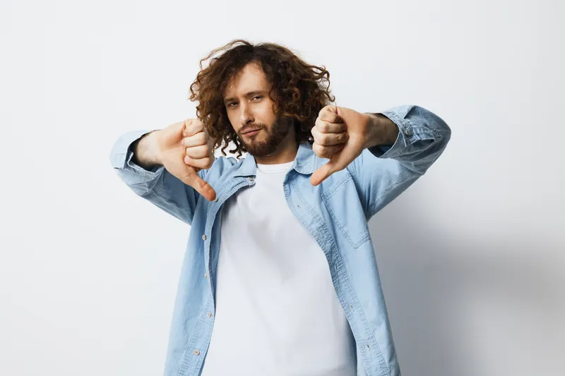 Young man with curly hair showing thumbs down, expressing dissatisfaction, dressed in casual clothing against a neutral background, conveying frustration and annoyance