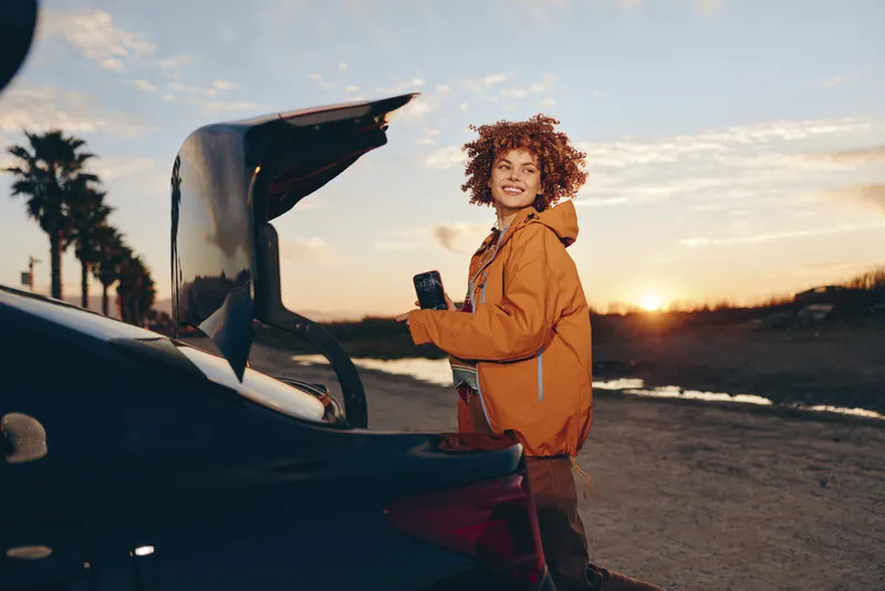 Woman lifestyle near open car trunk at sunset, curly hair portrait, holding smartphone, relaxed smile, warm golden light, casual jacket, outdoors travel mood, soft breeze, natural colors