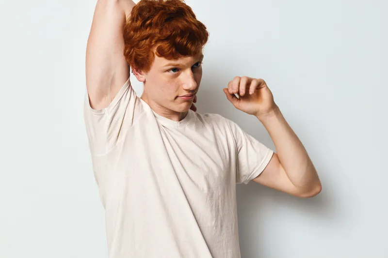 Young man with red hair wearing light t shirt stretching and lifting arm on white background for health care and personal hygiene concept.