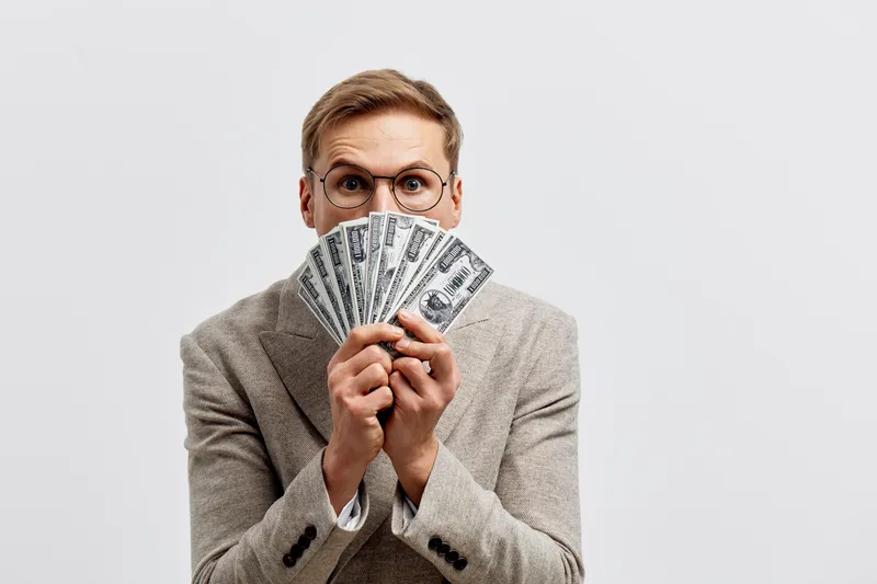 man with glasses holding US dollar bills in hands wear beige suit excited expression isolated on light background finance money concept