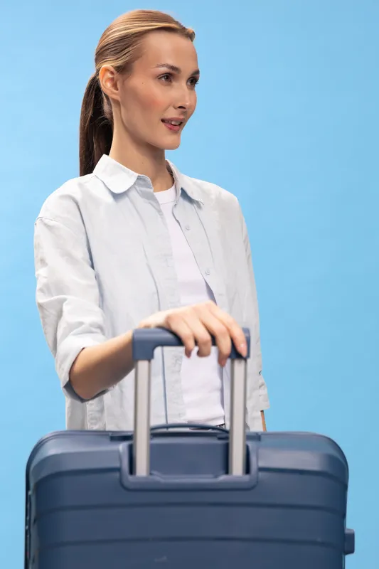 Young woman with ponytail wearing casual shirt holding blue suitcase handle standing against light blue background. Concept for travel, journey, vacation, and tourism with relaxed mood.