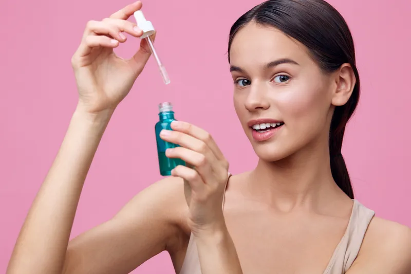 Young woman applying facial serum on a pink background, showcasing beauty and skincare routine with a smile, ideal for healthy skin concepts