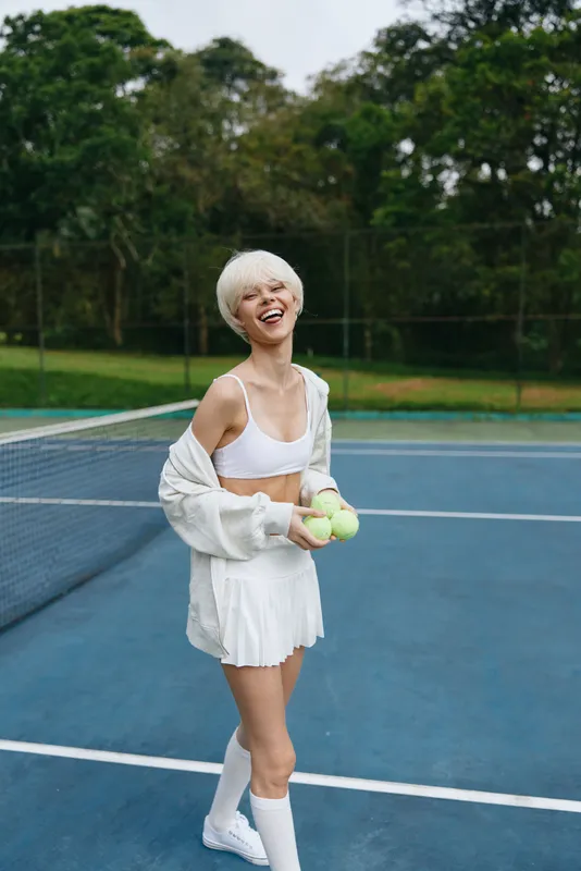 Cheerful young woman enjoying tennis on a sunny day in a white outfit with tennis balls on a court surrounded by green foliage
