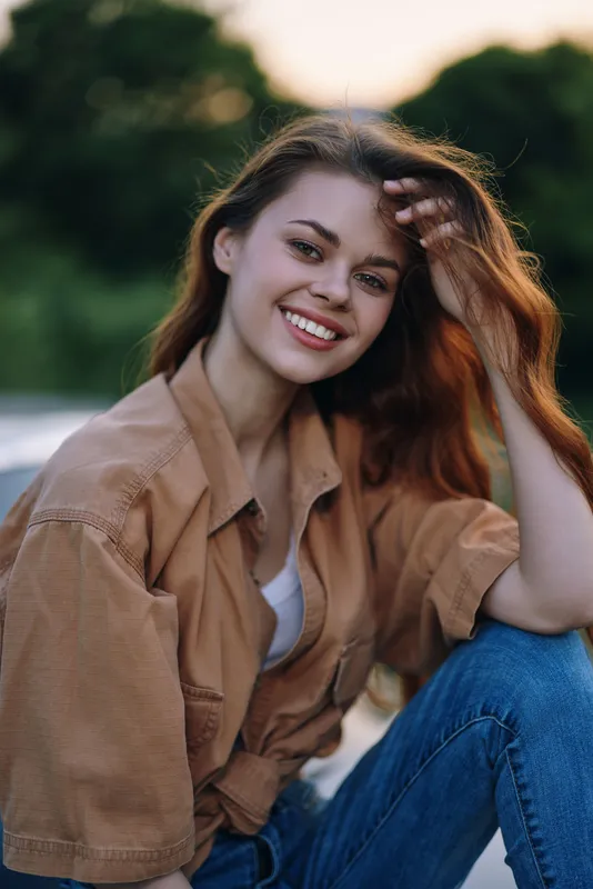 Smiling young woman with long hair in casual outfit sitting outdoors, conveying joy and confidence in a natural setting, perfect for lifestyle and beauty themes.