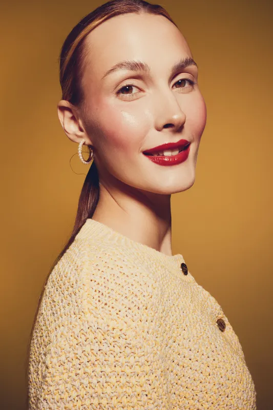 Young woman with red lipstick and hoop earrings smiles confidently wearing a yellow knitted sweater. Studio portrait on warm brown background with soft lighting and natural makeup.