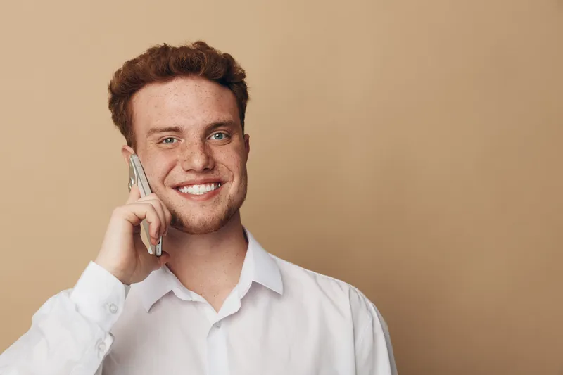 Smiling young man with curly hair talks on smartphone wearing white shirt. Isolated on beige background. Concept of communication, technology, and positive emotions.