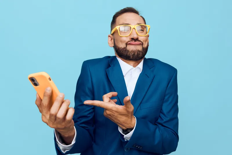 A business professional stands against a solid blue background, isolated and vibrant. The man exudes confidence, pride, and approachability, as he holds a phone and makes a bold gesture.