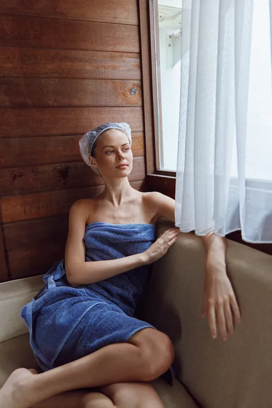 Relaxed woman wrapped in blue towel with shower cap, sitting comfortably on beige sofa near window, enjoying peaceful natural light in wooden interior setting.