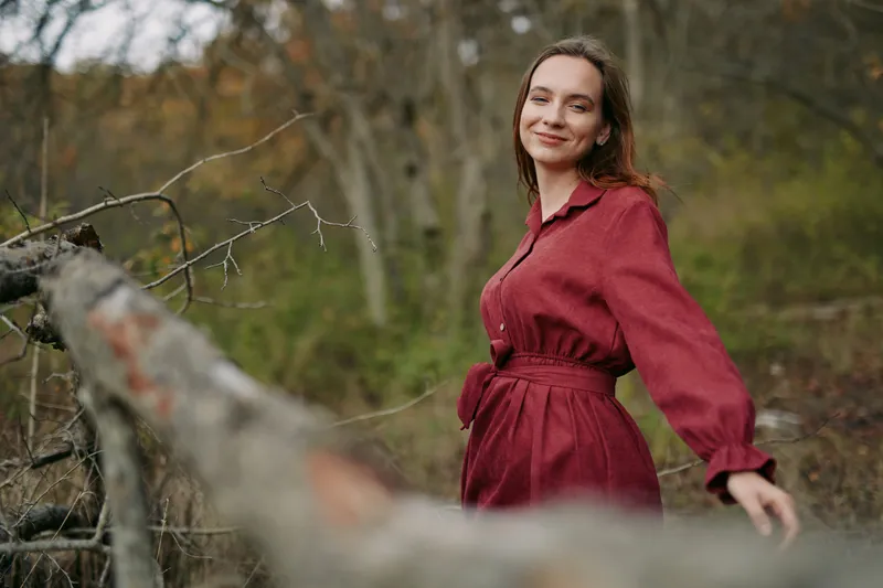Authentic, genuine moment in nature as a woman in a belted red dress stands by a rustic railing, conveying calm, warmth, and sincere presence.