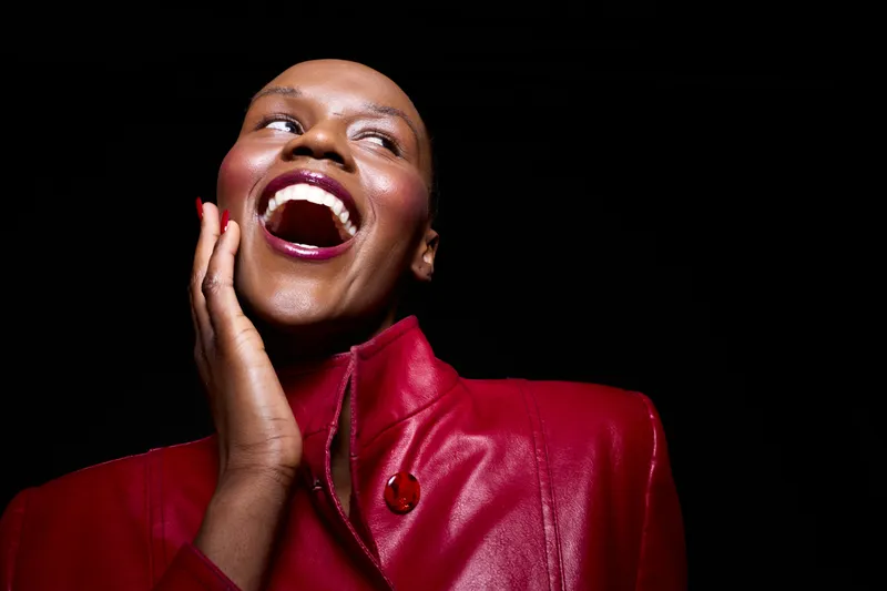 Happy African woman with dark skin laughs joyfully wearing red leather jacket, isolated on black background. Concept of beauty, fashion, confidence, and positive emotion.