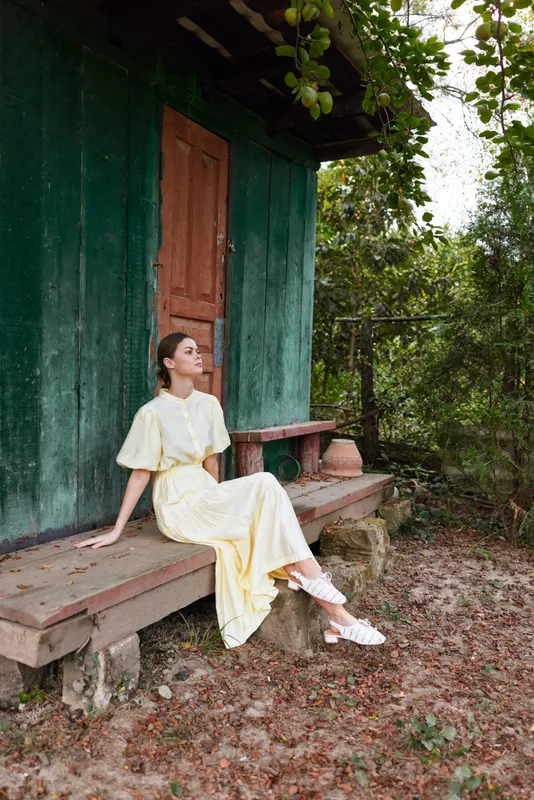 A woman in a pale yellow dress sits on a wooden bench outside a rustic green house, enjoying a tranquil moment in a sunny garden, with soft light and calm atmosphere.