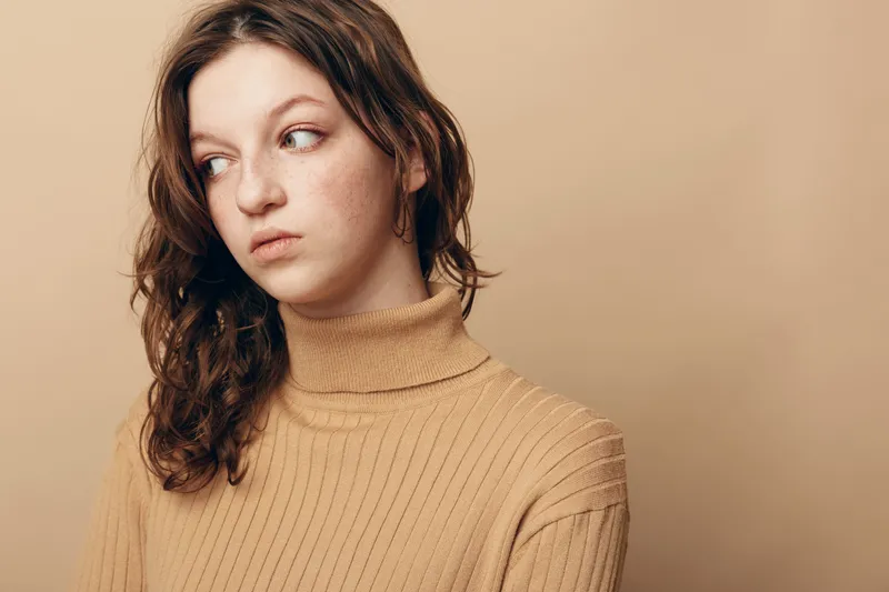 Young woman with curly brown hair wearing beige turtleneck looks away with thoughtful expression on neutral background. Concept of contemplation, mood, and casual fashion portrait.