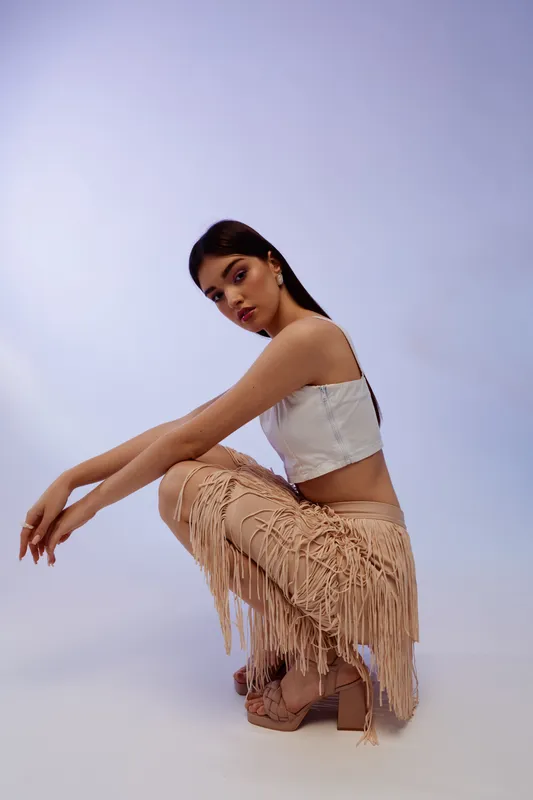 Young woman in fashion outfit poses squatting on a light background. She wears a white crop top and fringed beige pants. Concept of beauty, style, and modern fashion.