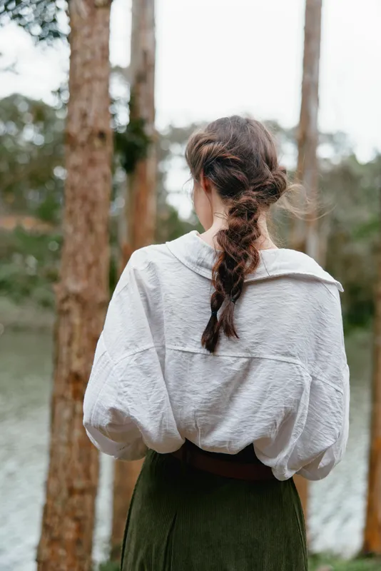 A woman with a braided hairstyle wearing a loose white blouse and green skirt, standing by a serene lake surrounded by lush trees, exuding a calm and natural vibe